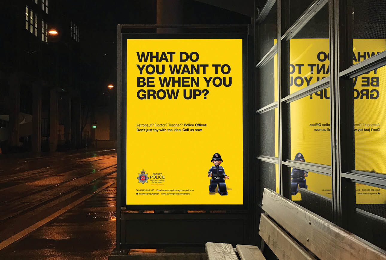 Surrey Police recruitment poster at bus stop with yellow background and toy officer figure at night